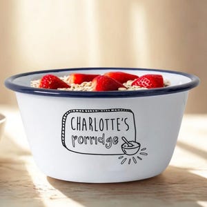 May include: A white enamel bowl with a blue rim, filled with porridge and sliced strawberries. The bowl features the text "CHARLOTTE'S porridge" in black. A smaller white bowl with strawberries is visible in the background.