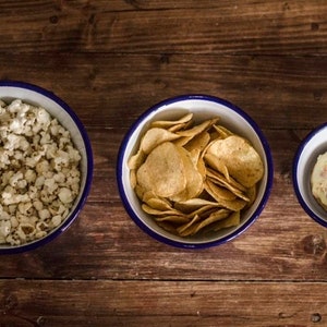 May include: Three white enamel bowls with blue rims, each filled with a different snack: popcorn, tortilla chips, and a frosted donut.