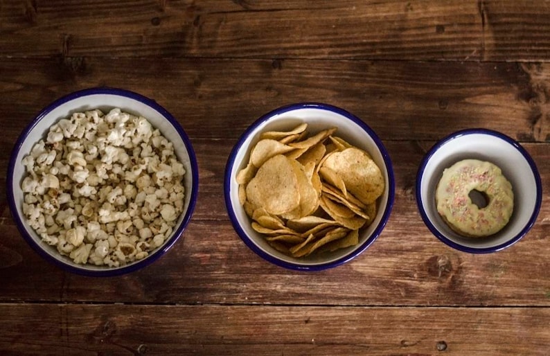 May include: Three white enamel bowls with blue rims, each filled with a different snack: popcorn, tortilla chips, and a frosted donut.