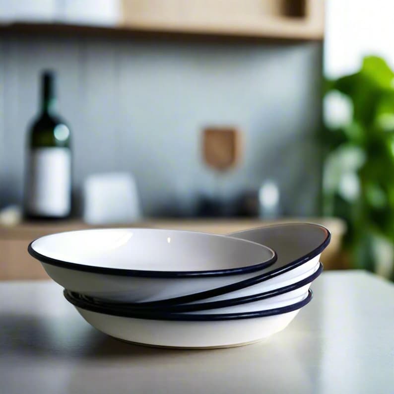May include: A stack of four white enamel bowls with black rims. The bowls are nested together and are sitting on a white surface.