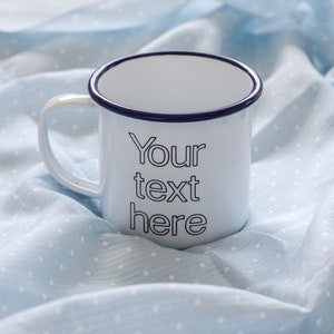 May include: White enamel mug with a blue rim and the text "Your text here" printed on the front. The mug is sitting on a light blue fabric with white polka dots.