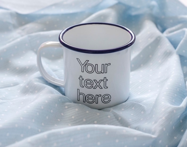 May include: White enamel mug with a blue rim and the text "Your text here" printed on the front. The mug is sitting on a light blue fabric with white polka dots.