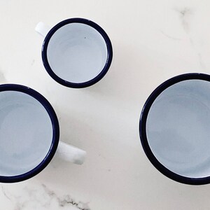 May include: Three enamel mugs with blue rims. The mugs are white on the inside and have a white background. The mugs are different sizes.