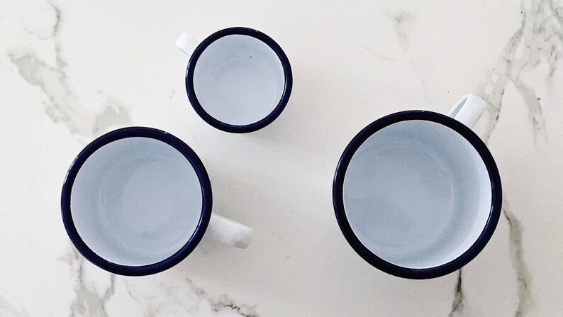 May include: Three enamel mugs with blue rims. The mugs are white on the inside and have a white background. The mugs are different sizes.