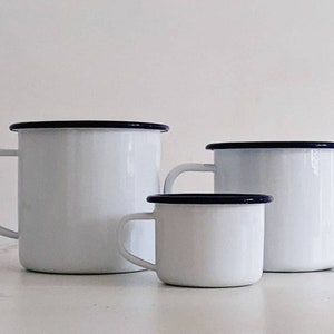 May include: Three white enamel mugs with blue rims. The mugs are of different sizes, with the largest mug in the back and the smallest mug in the front. The mugs are on a white marble surface.
