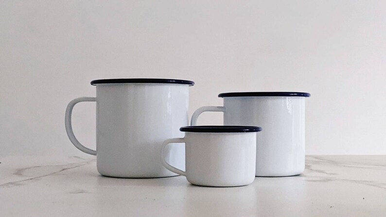 May include: Three white enamel mugs with blue rims. The mugs are of different sizes, with the largest mug in the back and the smallest mug in the front. The mugs are on a white marble surface.