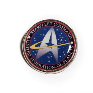 Puede incluir: Un pin de plata con un logotipo de Starfleet Command azul, rojo y amarillo. El logotipo presenta una estrella blanca con una luna creciente amarilla y el texto "United Federation of Planets" y "Starfleet Command".