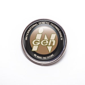 May include: A silver pin with a black circle containing the text "International Genetic Technologies" and the logo "iGen" in white and green letters on a brown background. The text "We Make The Future" is printed below the logo.