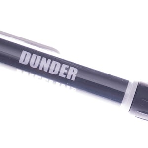 Puede incluir: Un bolígrafo gris y blanco con la palabra "DUNDER MIFFLIN" impresa en el lateral.