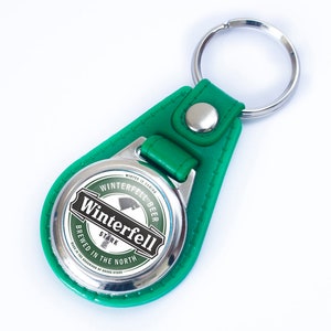 Puede incluir: Llavero de cuero verde con un círculo de metal plateado que presenta una etiqueta que dice "Winterfell Beer, Stark, Brewed in the North". La etiqueta tiene una imagen en blanco y negro de un lobo.