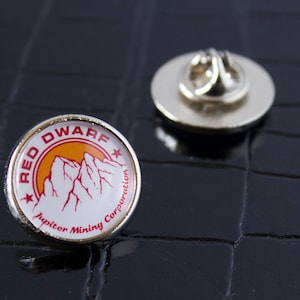 May include: A silver pin with a white background and a red and orange logo that says "Red Dwarf Jupiter Mining Corporation".