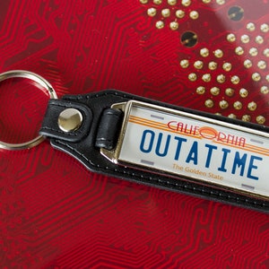 Könnte beinhalten: Schwarzer Leder-Schlüsselanhänger mit einer silbernen Metallplatte, die ein stilisiertes kalifornisches Nummernschild mit dem Text "OUTATIME" und "The Golden State" zeigt.