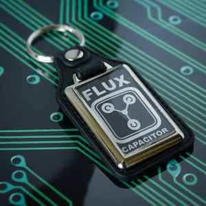 Puede incluir: Un llavero de cuero negro con un dije de metal plateado. El dije tiene las palabras "Flux Capacitor" y una imagen estilizada de una placa de circuito.