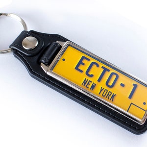 Może przedstawiać: Czarny skórzany brelok do kluczy z żółtym metalowym brelokiem w kształcie tablicy rejestracyjnej z napisem "ECTO-1" i "NEW YORK" poniżej.
