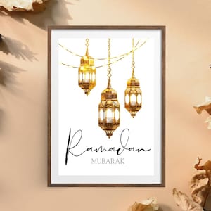 Puede incluir: Impresión enmarcada con tres faroles dorados ornamentados suspendidos de una cadena dorada sobre un fondo blanco. La palabra "Ramadán" está escrita en una elegante escritura, con "MUBARAK" debajo.