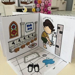 May include: A paper craft book featuring a Wudu area illustration with a stained glass window, a sign reading Wudu Area, and a cartoon character washing feet at a faucet. The floor shows a tiled pattern with a mat, a pair of sandals, and a blue water puddle.
