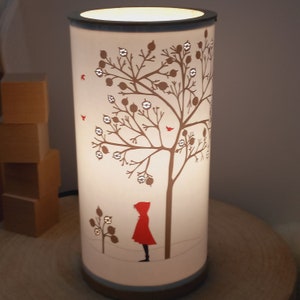 Op de afbeelding: Cilindrische lamp met een warme gloed, met een bruin boomontwerp met bessen en rode vogels. Een silhouet van een persoon in een rode cape staat onder de boom. De lamp heeft een donkere rand en basis.