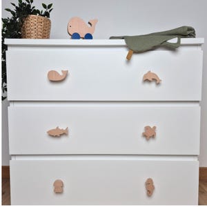 Puede incluir: Una cómoda blanca con tiradores de madera con forma de criaturas marinas. Los tiradores incluyen una ballena, un delfín, un tiburón, una tortuga, una medusa y un caballito de mar. Un juguete de ballena de madera con ruedas azules está encima.