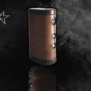 May include: A brown leather vape mod with three buttons and a black trim. The mod is sitting on a black surface with smoke in the background.