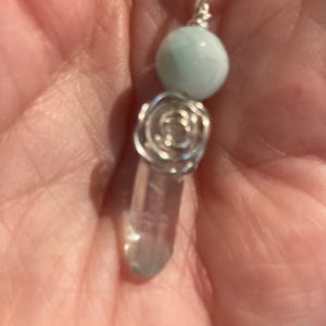 May include: A handmade pendant necklace featuring a clear quartz crystal, a light blue bead, and a silver wire rose design. The pendant hangs from a black cord, creating a unique and elegant accessory.