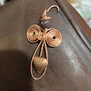 Puede incluir: Colgante artesanal de alambre de cobre con un dije en forma de corazón. El colgante presenta alas de alambre enrollado, una cuenta morada oscura y un corazón de cobre. El diseño es intrincado y detallado, mostrando la habilidad del artesano.