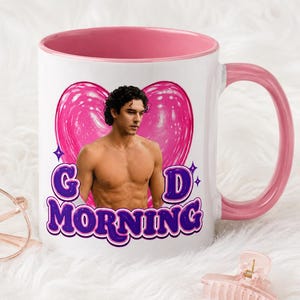 Puede incluir: Una taza de cerámica blanca con interior y asa de color rosa. El frente presenta un gráfico de una persona con cabello oscuro y rizado, un fondo de corazón rosa grande y el texto morado "GOOD MORNING" en una fuente retro, negrita y redondeada.