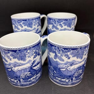 以下が含まれることがあります： 青と白の転写プリントが施された陶器製マグカップ4個セット。雪に覆われた冬の風景、家、木々、人物が描かれており、上部の縁にはヒイラギの葉と実の装飾的なボーダーがあしらわれています。