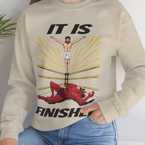 Op de afbeelding: Beige sweatshirt met een afbeelding van een figuur in een boksring, met de tekst "IT IS FINISHED". De afbeelding toont een figuur met uitgestrekte armen boven een verslagen rode figuur. De sweatshirt heeft lange mouwen en een ronde hals.
