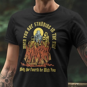 May include: Black t-shirt with a vintage-style illustration of a group of people standing in a fire. The text "When you are standing in the fire" is written in a circle around the image. Below the image, the text "May the fourth be with you" is written in a smaller font.