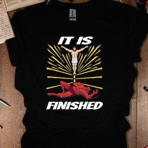 Könnte beinhalten: Schwarzes T-Shirt mit einem grafischen Design, das eine Figur mit ausgestreckten Armen über einer roten Figur zeigt, mit den Worten "IT IS FINISHED" in Weiß. Das Design ist auf dem T-Shirt zentriert.