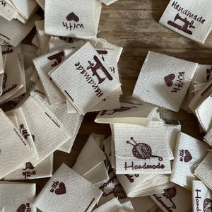 May include: A collection of off-white fabric tags scattered on a wooden surface. Each tag features the word "Handmade" in brown script, along with a small illustration of a sewing machine, yarn, or a heart. The tags are small, square, and appear to be for handmade crafts.