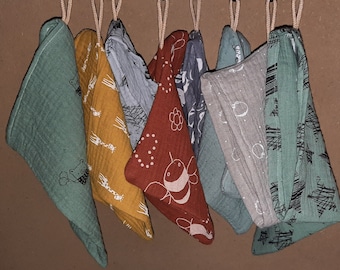 Puede incluir: Una colección de siete baberos triangulares para bebés, colgados de una cuerda. Los baberos presentan varios diseños, incluyendo abejas, osos y diseños abstractos, en tonos verdes, amarillos, rojos y grises. Están hechos de tela suave.