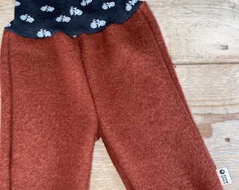 May include: Rust-colored pants featuring a textured fabric, with dark gray cuffs and a waistband patterned with small white bicycle icons. A small white tag on the side seam displays the text "HAND MADE" in black lettering.