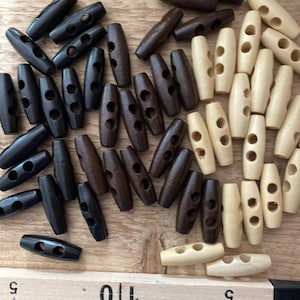 May include: A collection of wooden toggle buttons in various shades of brown and natural wood. The buttons are oblong with two holes for sewing. The buttons are arranged on a wooden surface.