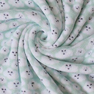 Puede incluir: Una manta suave y afelpada con un patr&oacute;n repetido de caras de foca blancas sobre un fondo verde claro. La manta est&aacute; doblada y arremolinada, mostrando la textura de la tela. Las focas tienen ojos y narices negras.