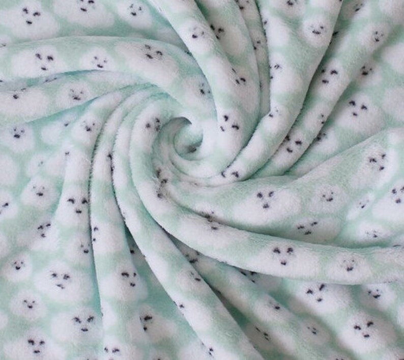 Puede incluir: Una manta suave y afelpada con un patr&oacute;n repetido de caras de foca blancas sobre un fondo verde claro. La manta est&aacute; doblada y arremolinada, mostrando la textura de la tela. Las focas tienen ojos y narices negras.