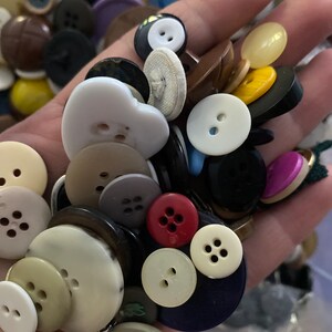 Puede incluir: Una colección de botones variados en diferentes tamaños, formas y colores, incluyendo blanco, negro, marrón, rojo y amarillo. Algunos botones tienen dos agujeros, mientras que otros tienen cuatro. Los botones se sostienen en una mano.