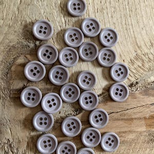 Op de afbeelding: Een verzameling lichtpaarse, ronde knopen met elk vier gaatjes. De knopen liggen verspreid op een houten oppervlak, wat hun uniforme kleur en ontwerp benadrukt. Ideaal voor naai- en knutselprojecten.