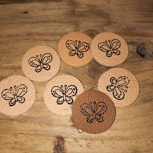 Könnte beinhalten: Sieben runde Lederaufnäher mit einem Schmetterlingsdesign. Die Aufnäher sind in verschiedenen Beige- und Brauntönen gehalten, mit dem Schmetterlingsdesign in schwarzer Tinte. Auf einer Holzoberfläche angeordnet.