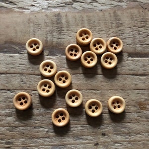 Op de afbeelding: Een verzameling kleine, ronde houten knopen met elk vier gaatjes. De knopen zijn lichtbruin van kleur en liggen op een verweerd houten oppervlak. Ideaal voor naai- en knutselprojecten.