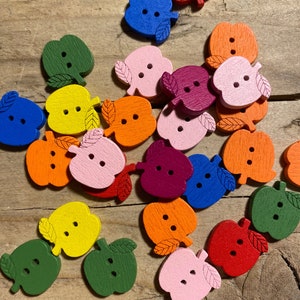 Könnte beinhalten: Eine Sammlung bunter, apfelförmiger Holzknöpfe in verschiedenen Farbtönen, darunter Rot, Orange, Gelb, Grün, Blau, Rosa und Lila. Jeder Knopf hat zwei Löcher und ein Blattdetail. Die Knöpfe liegen auf einer Holzoberfläche verstreut.