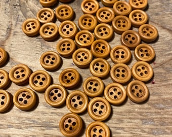 May include: A selection of small, round wooden buttons, each with four holes. The buttons are a light brown hue and are arranged on a wooden surface. These buttons are suitable for sewing and craft activities.