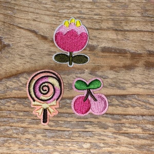 Könnte beinhalten: Drei gestickte Aufnäher: eine rosa Blume mit gelben Akzenten, ein Wirbellutscher in Rosa und Pfirsich und ein Paar rosa Kirschen mit grünen Stielen. Die Aufnäher befinden sich auf einer Holzoberfläche.