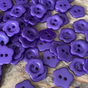 Puede incluir: Una colección de botones morados vibrantes con forma de flor. Cada botón tiene dos agujeros y un acabado brillante. Estos botones decorativos están dispersos sobre una superficie de madera, ideales para coser y manualidades.