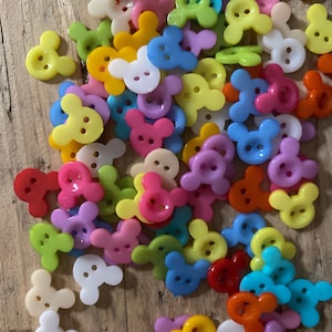 Può includere: Una collezione di bottoni colorati in plastica a forma di testa di topo. I bottoni sono in vari colori, tra cui rosso, rosa, giallo, blu, verde e bianco. I bottoni sono sparsi su una superficie di legno.