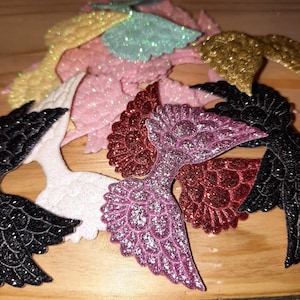 Könnte beinhalten: Eine Sammlung glitzernder, flügelförmiger Verzierungen in verschiedenen Farben, darunter Schwarz, Weiß, Rosa, Rot, Gold und Türkis. Die Flügel sind auf einer Holzoberfläche angeordnet und zeigen ihre glitzernde Textur und ihr detailliertes Design.