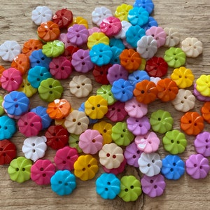 Puede incluir: Una colección de botones coloridos con forma de flor en varios tonos, incluyendo blanco, rojo, naranja, amarillo, verde, azul y morado. Los botones son de plástico y tienen dos agujeros para coser. Están dispersos sobre una superficie de madera.