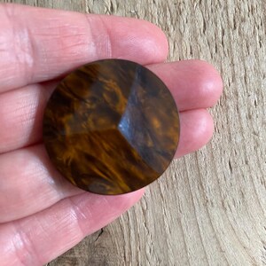Op de afbeelding: Een grote, ronde, bruine knoop met een facetontwerp. De knoop heeft een gemarmerd uiterlijk met tinten amber en donkerbruin. De knoop wordt in een hand gehouden, wat de grootte en details laat zien. Deze knoop is geschikt voor naaiprojecten.