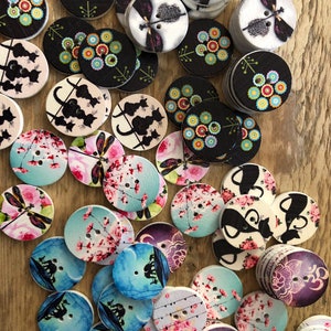 Könnte beinhalten: Eine Sammlung von Holzknöpfen mit verschiedenen Designs. Einige Knöpfe zeigen florale Muster in Schwarz, Weiß und bunten Farben. Andere Knöpfe zeigen Katzen-Silhouetten, Libellen und Kirschblüten. Die Knöpfe sind rund und haben zwei Löcher zum Annähen.