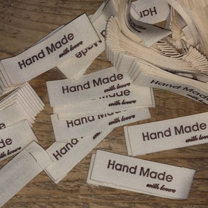 Könnte beinhalten: Cremefarbene Stoffetiketten mit dem Aufdruck "Hand Made with love" in Braun. Die rechteckigen Etiketten liegen auf einer Holzoberfläche, einige ordentlich gestapelt, andere verstreut.